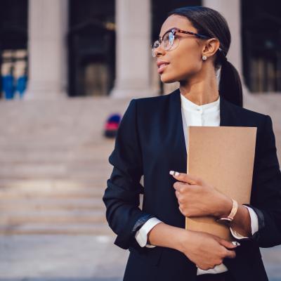 A young woman wearing glasses and business attire is standing outside a court house holding files and papers