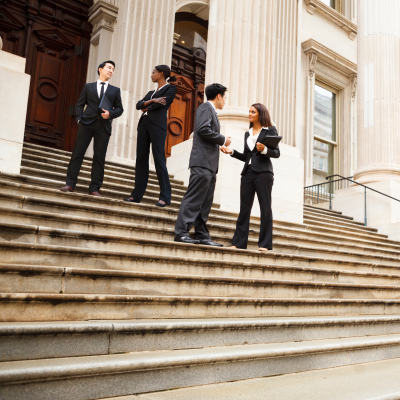 Diverse group of four adults in business suits talking while standing on the steps outside a large white building with large columns. Possibly a courthouse or an official government building.