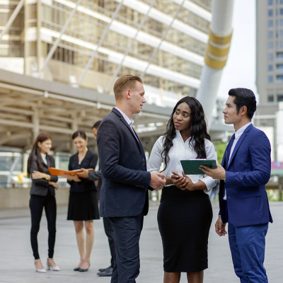 Diverse group of adults in business attire talking while standing outside a large modern office building with windows.