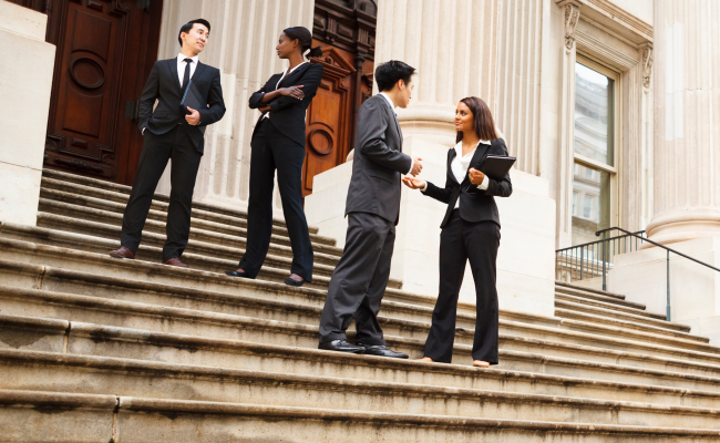 Diverse group of four adults in business suits talking while standing on the steps outside a large white building with large columns. Possibly a courthouse or an official government building.